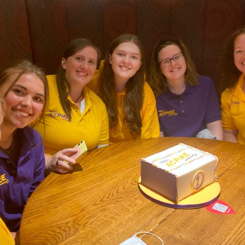 Smiling Staff in their colourful uniforms sat around a cake celebrating 1 year of Inspire to Aspire