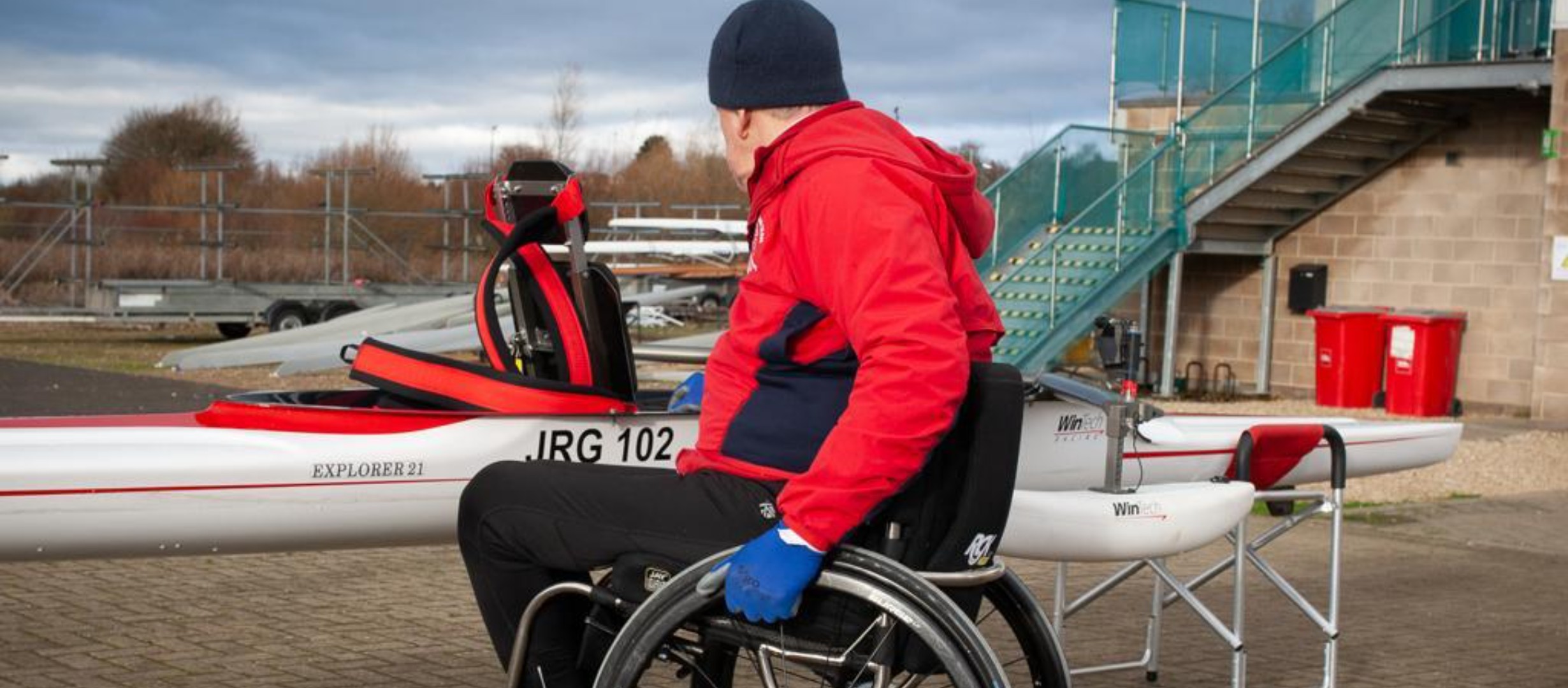 Rowing Boat on land with a rower in a red coat looking at it in his wheelchair. He is about to go on the water and is checking his equipment