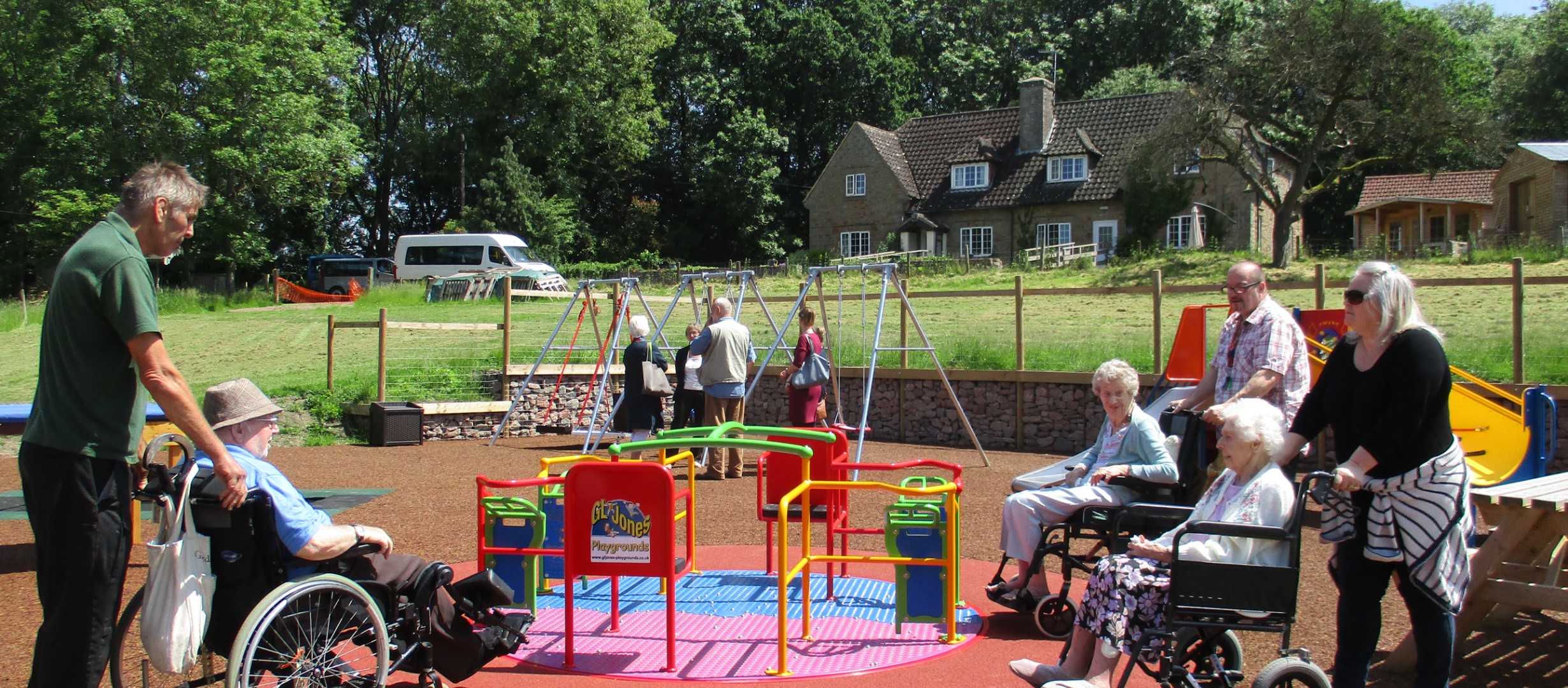 Image shows the Orchard Trust activity park with three people in wheelchairs and their carers about to use the roundabout. The orchard Trust orchards Care Home is in the background