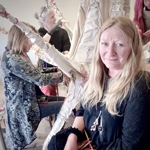 Lou, a white woman with fair hair, looks at the camera, while in the background Aly and Juliet work on a large, spikey, cream-coloured fabric sculpture