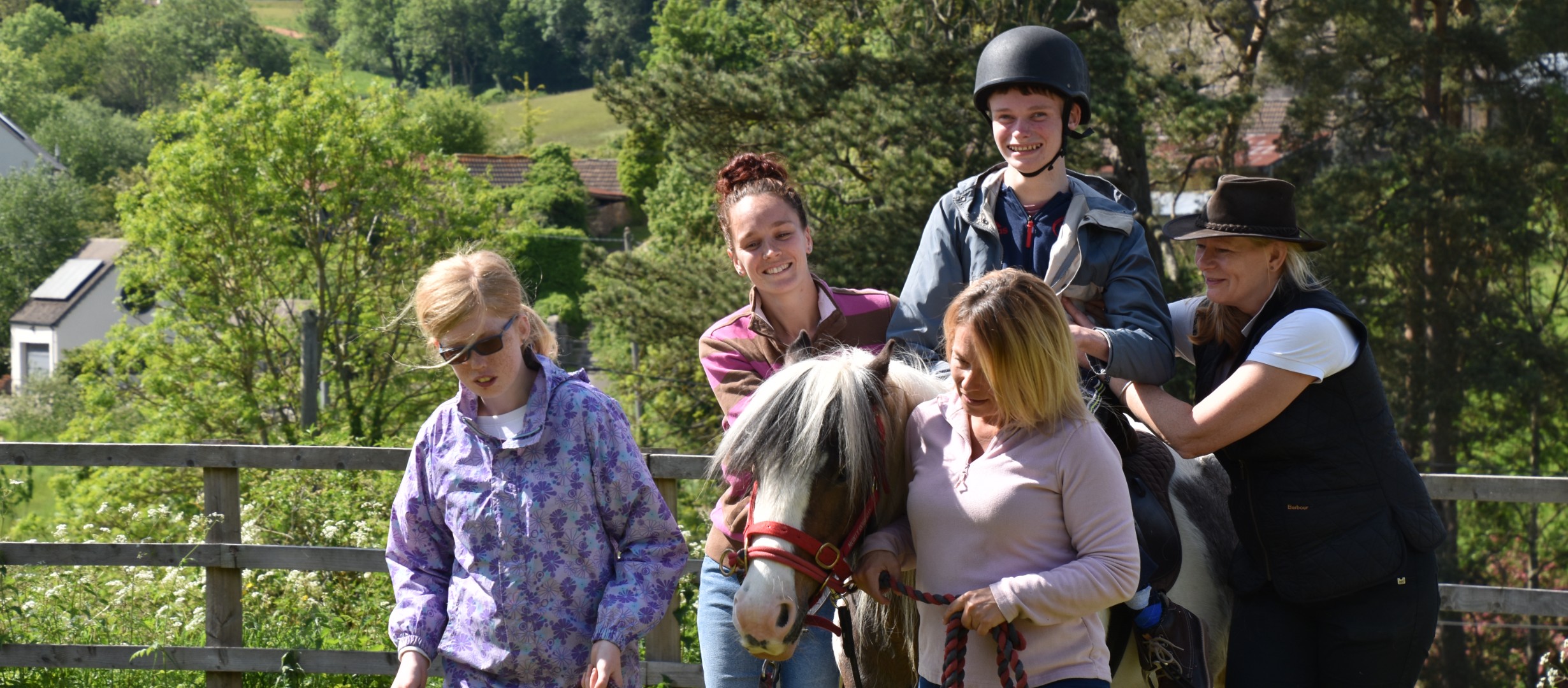 A young person riding a horse supported by three adults walking alongside the horse. The horse is being led by a young person