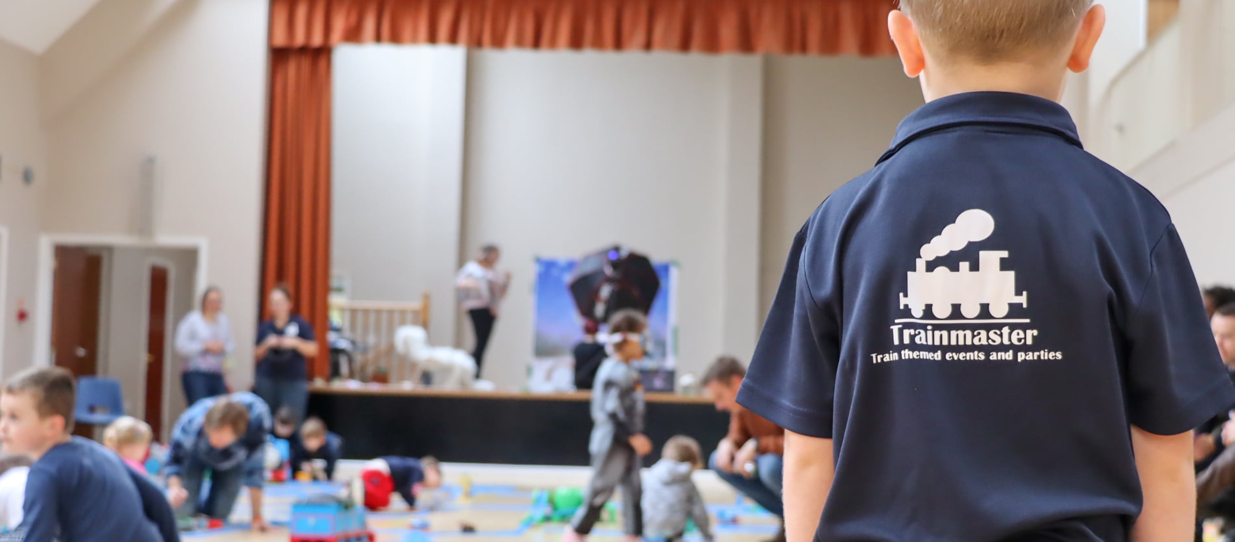 A child stands with his back to the camera. The Trainmaster Gloucestershire logo is seen on the back of his shirt. He faces a hall filled with train track and other train activities with children and adults playing freely