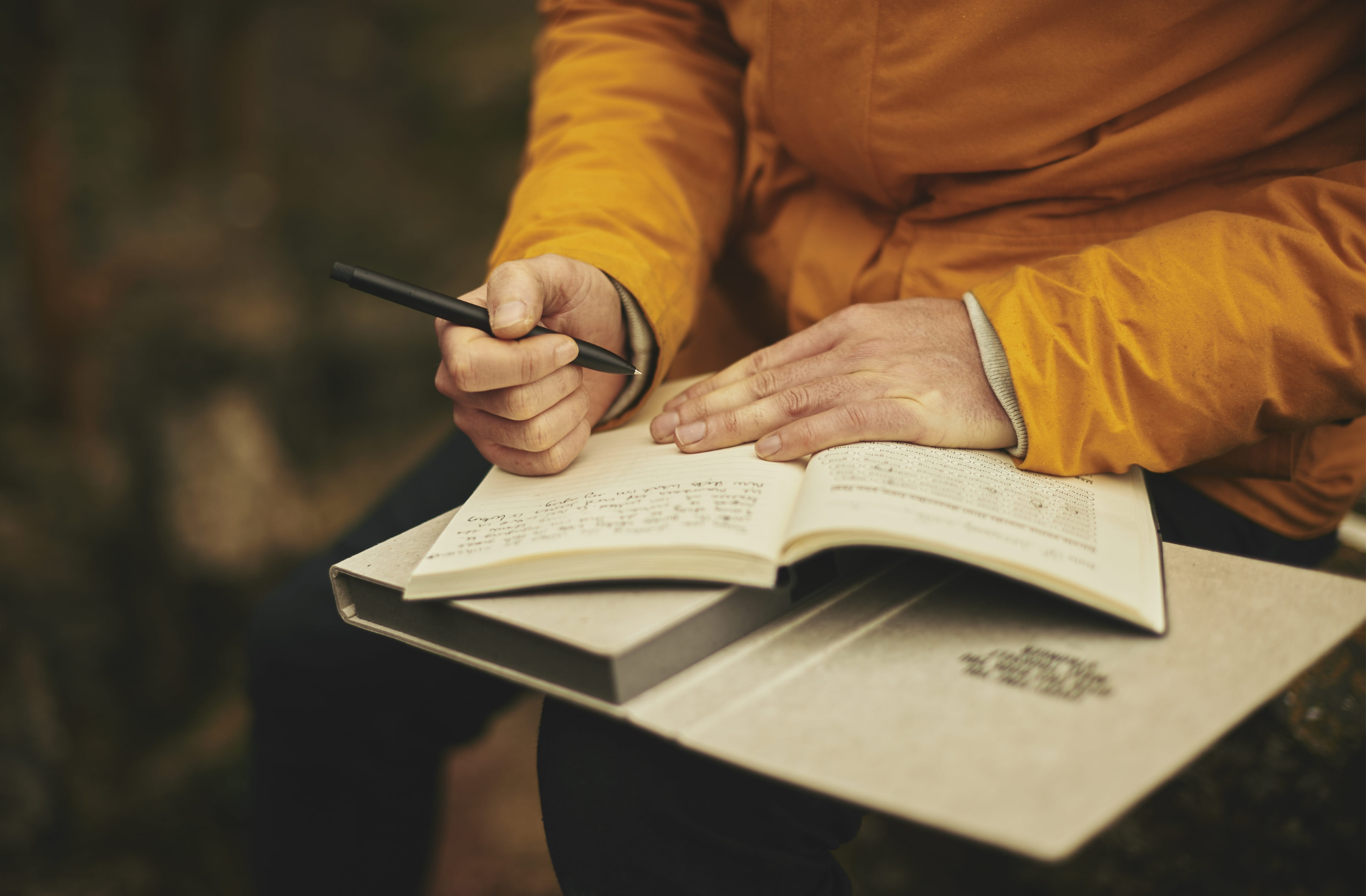 This is a photograph of an individual in a yellow jumper writing outside in a notebook. They are holding a pen in their left hand, and the notebook is spread open in their lap. The colours are soft oranges and very autumnal