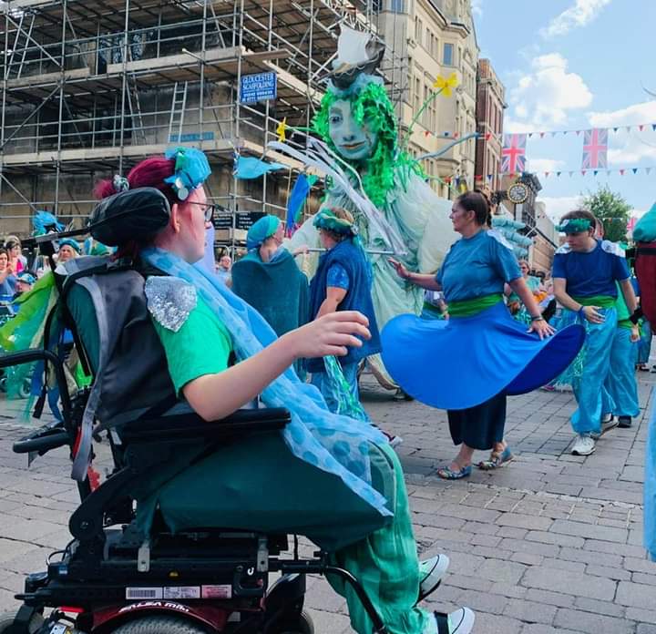 A photograph of participants from the Showtime drama group. There is a wheelchair user in the front of the image. Everyone is dressed in colourful blue and green outfits that represent water and rivers. In the background is a giant puppet of Sabrina the goddess of the river wye. This performance was for Hi!Street fest