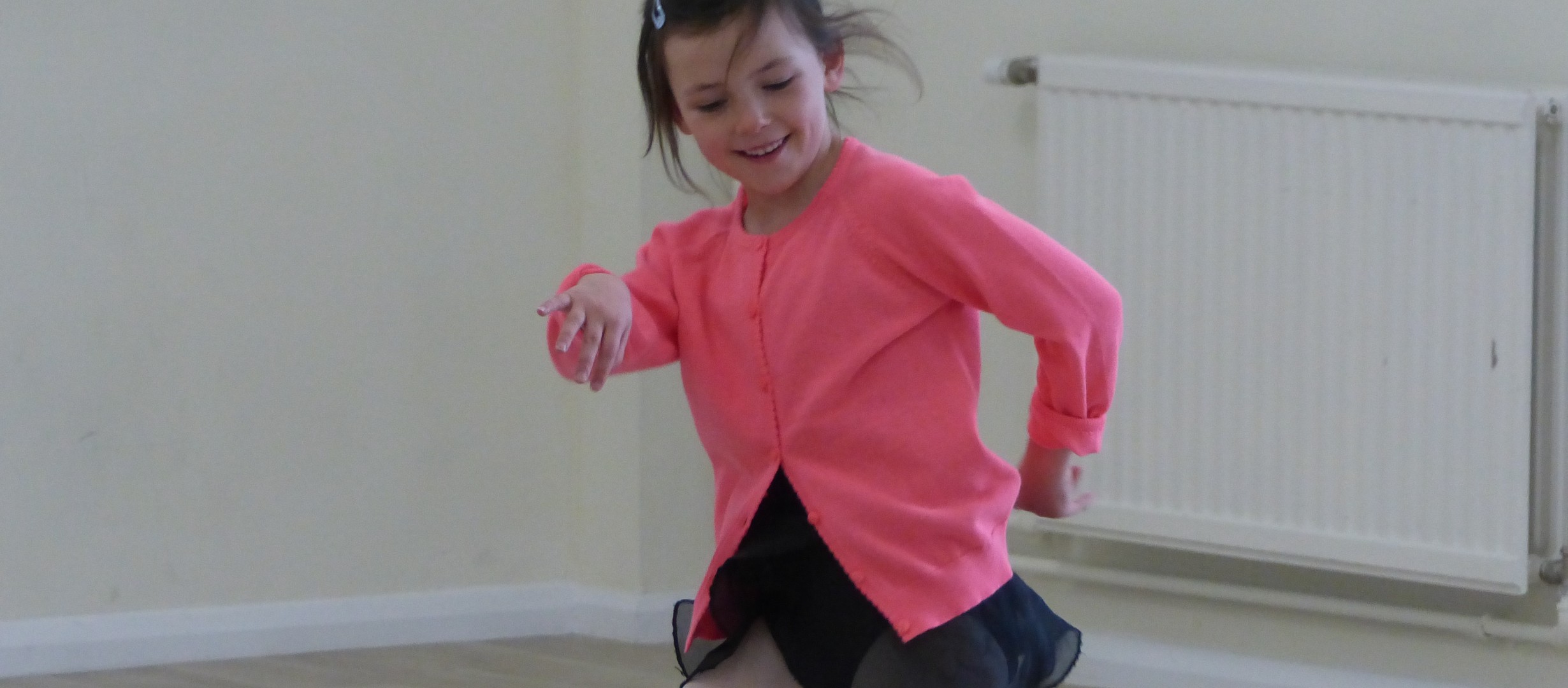 A female dancer aged about 8 years old with brown hair tied back smiling whilst jumping to the side. Wearing a pink cardigan and black ballet skirt.