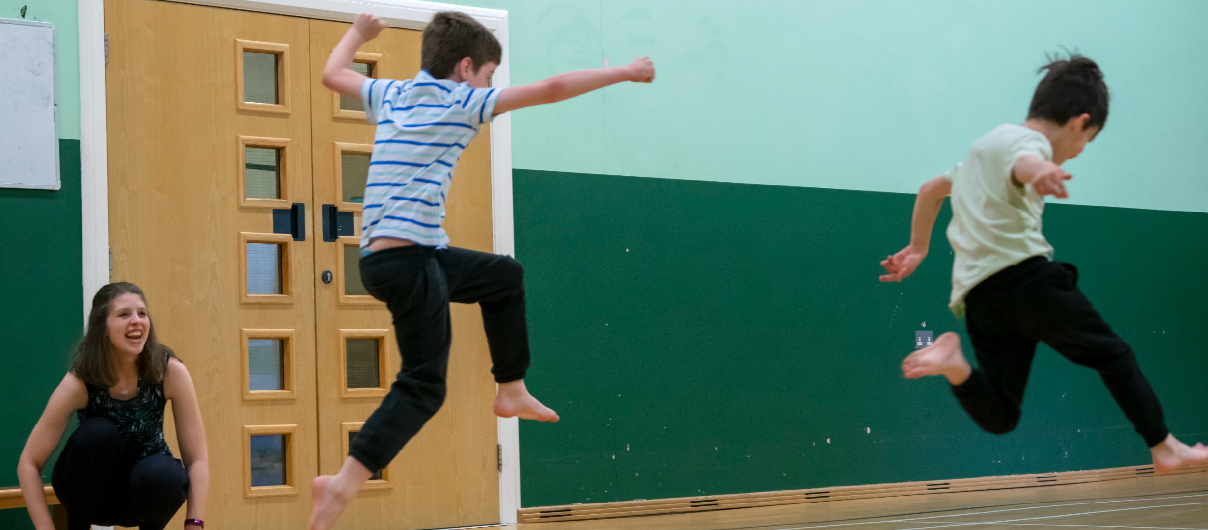 Photograph of two boys leaping over a stretch of blue material. Teacher, Katharine, in the background crouched down smiling at them!