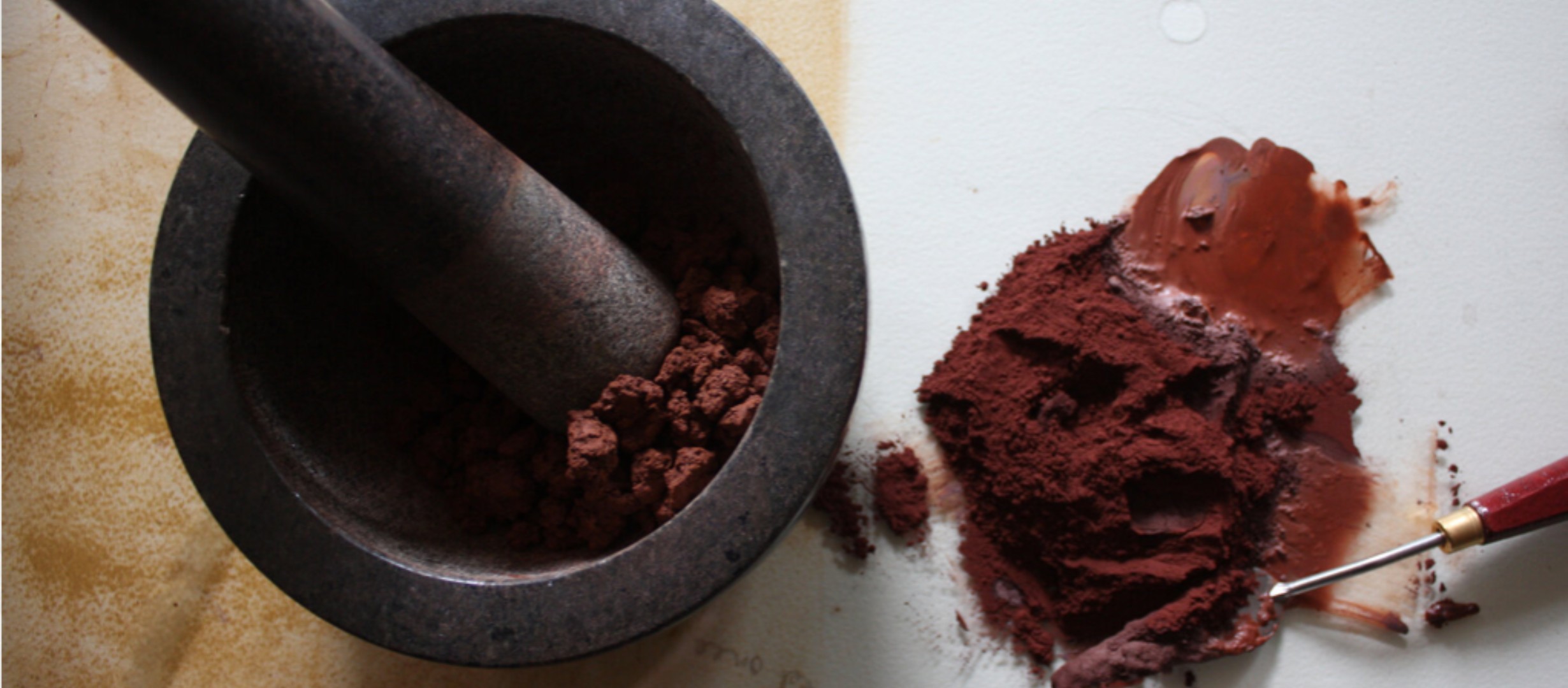A photograph of a pestle and mortor grinding brown earth pigments up, on a table with a pile of brown earth pigments on top