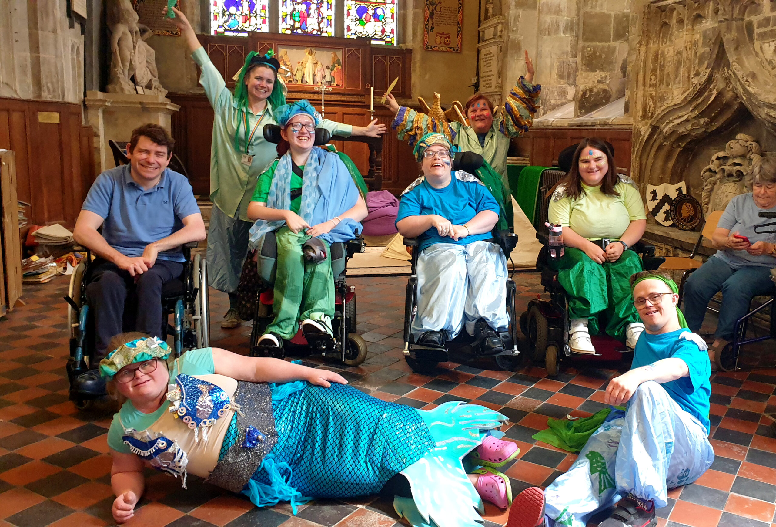 A photograph of members of the VIA Dance Company inside Gloucester cathedral. Participants are dressed in colourful blue outfits that represent water. There are 4 wheelchair users in the middle row. The particiants are in costume ready for Hi!Street fest, a summer festival in Gloucester city in 2023