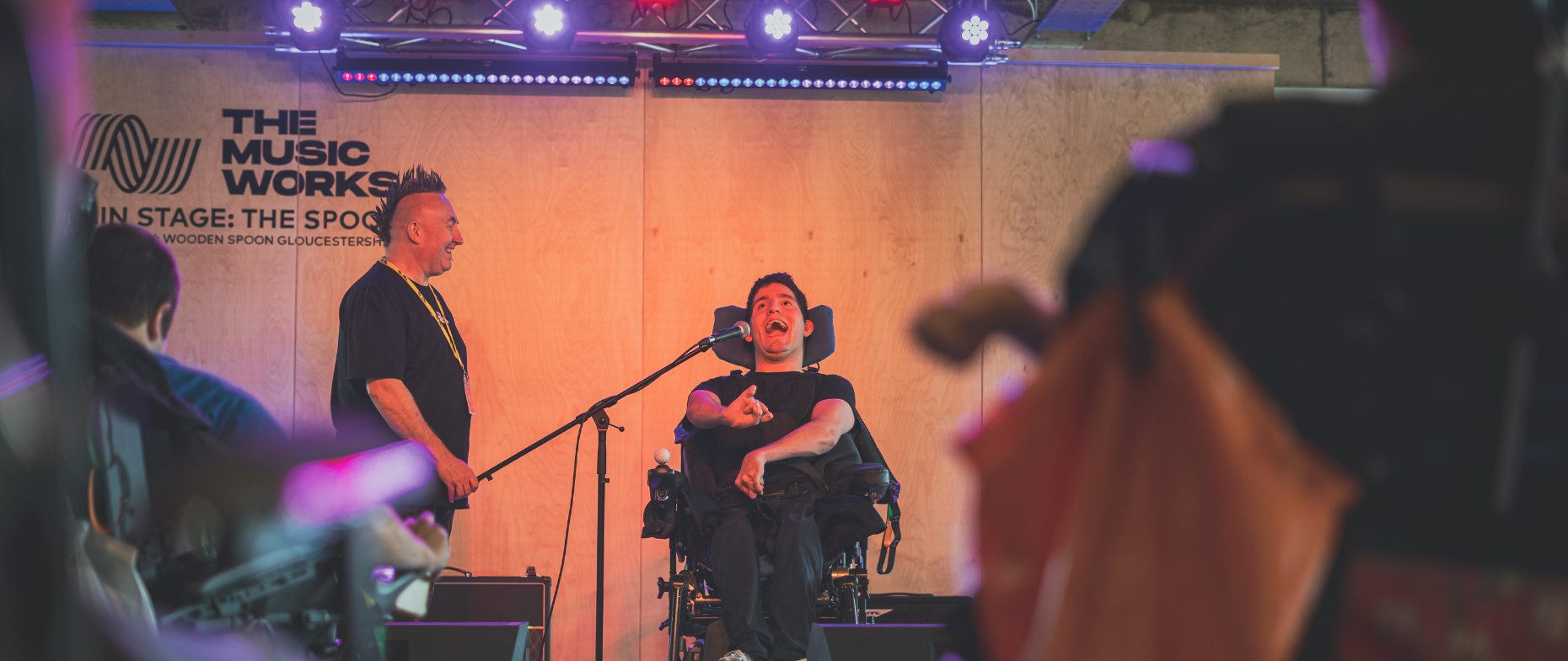 The Music Works' disability lead Lee Holder on stage with a young disabled performer singing.