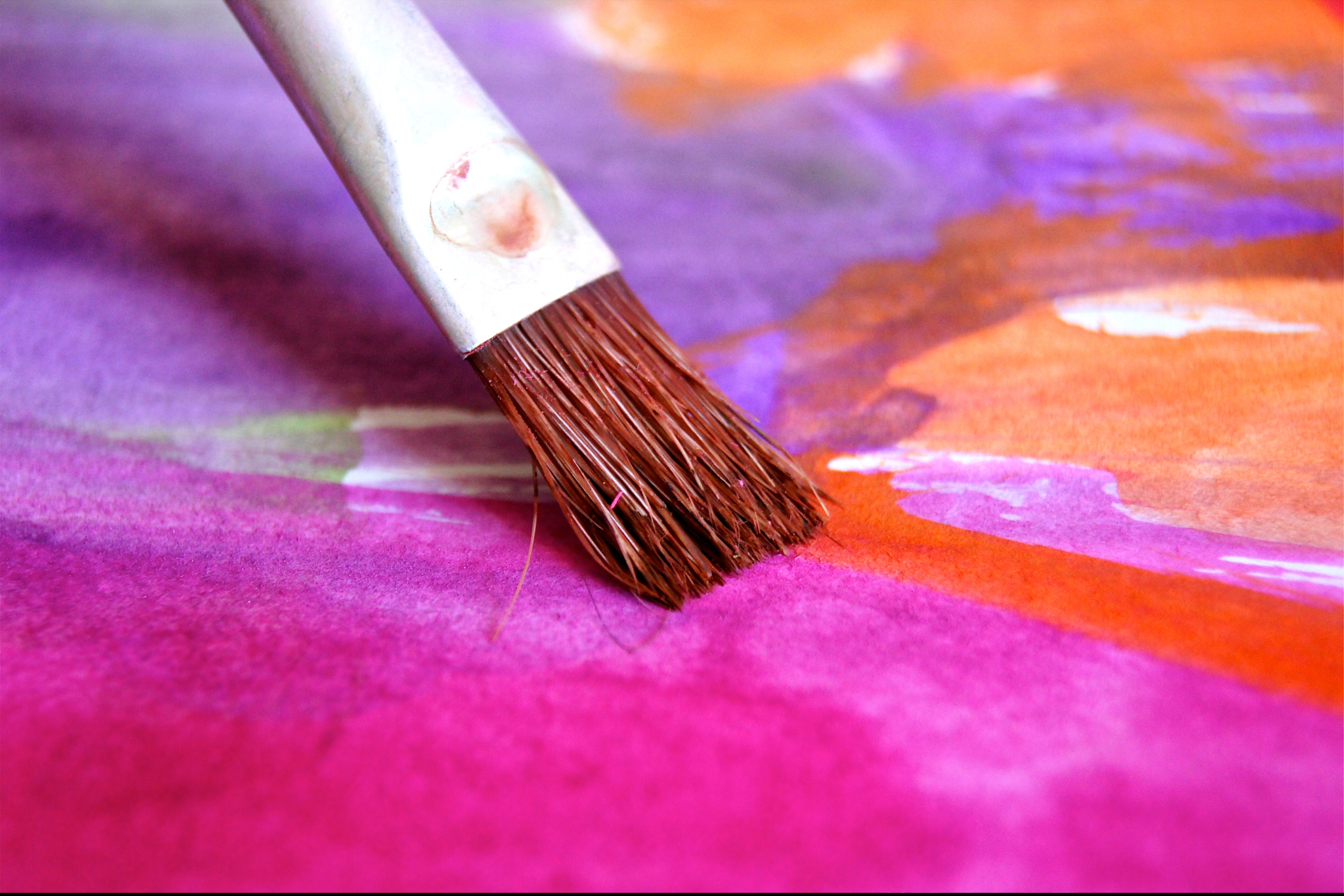 A photograph of a paintbrush sweeping across a vibrant purple, pink and orange painting. The paintbrush has brown bristles with a silver cap