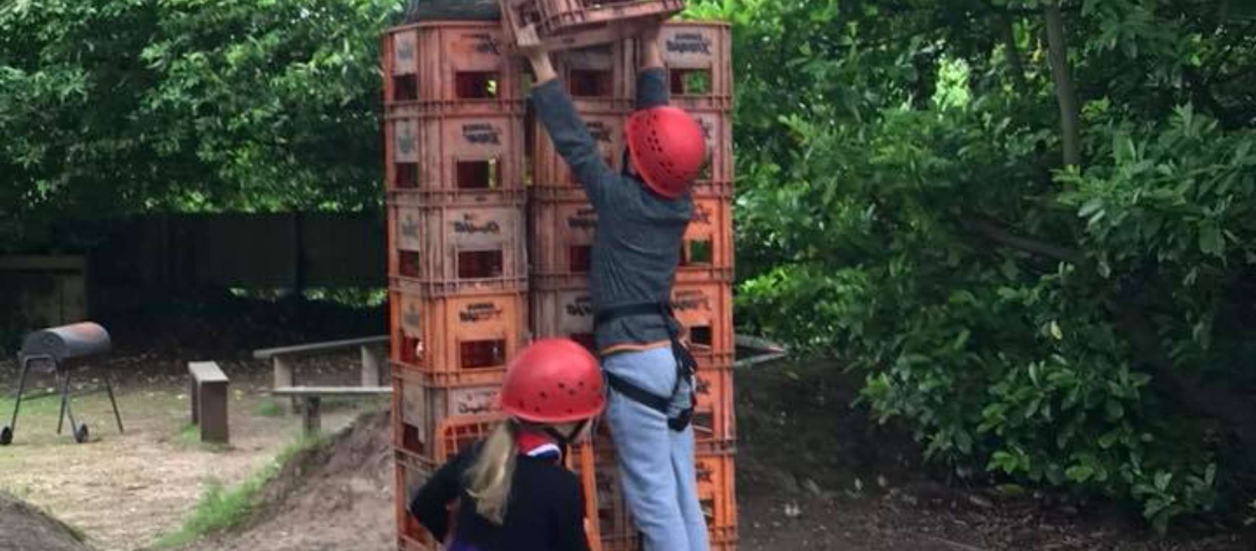 Two children wearing a harness and helmet stood in front of a stack of crates. One is handing a crate up in the air towards the top of the stack.