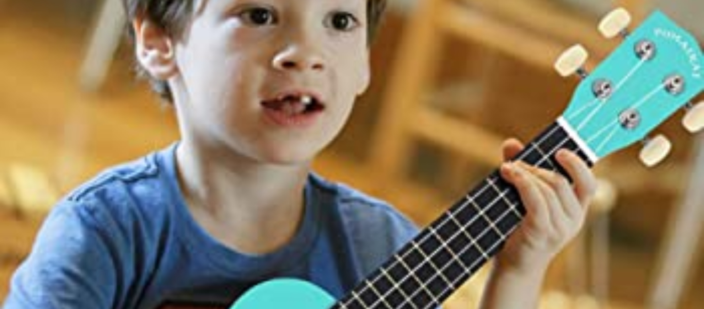 Primary age boy with dark tossled hair and missing teeth, holding an aqua ukulele. He’s sat cross legged on the floor with a look of concentration on his face. 