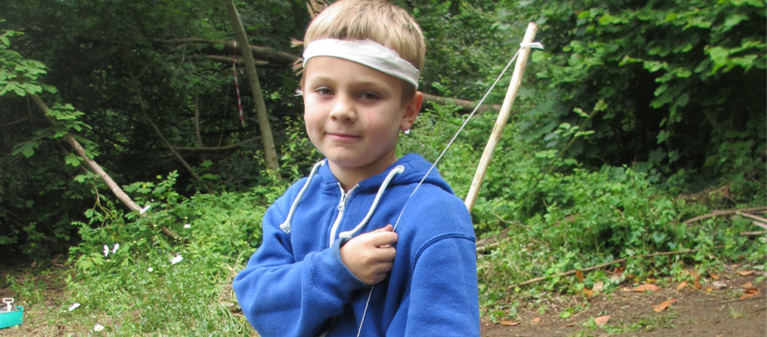 Child with short hair is stood wearing a blue hoody and white bandana holding a bow over their shoulder. Stood in a wooded clearing.