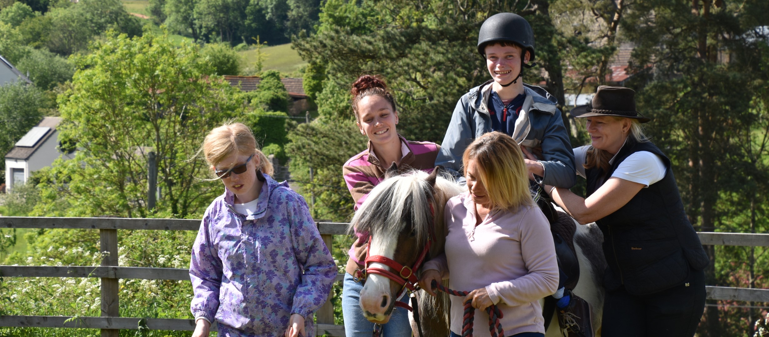 One person sat on a horse with two adults supporting them alongside the horse and two people leading the horse on reins. The background is a tree lined fence. 