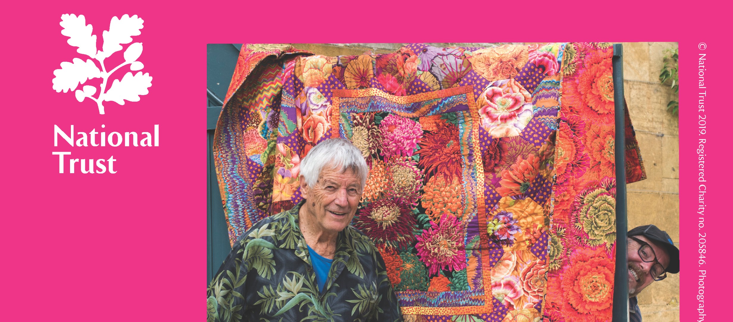 A bright pink background, to the left is the white oak leaf logo of the National Trust, to the right pictured is Kaffe Fassett standing in front of one of his colourful quilts.