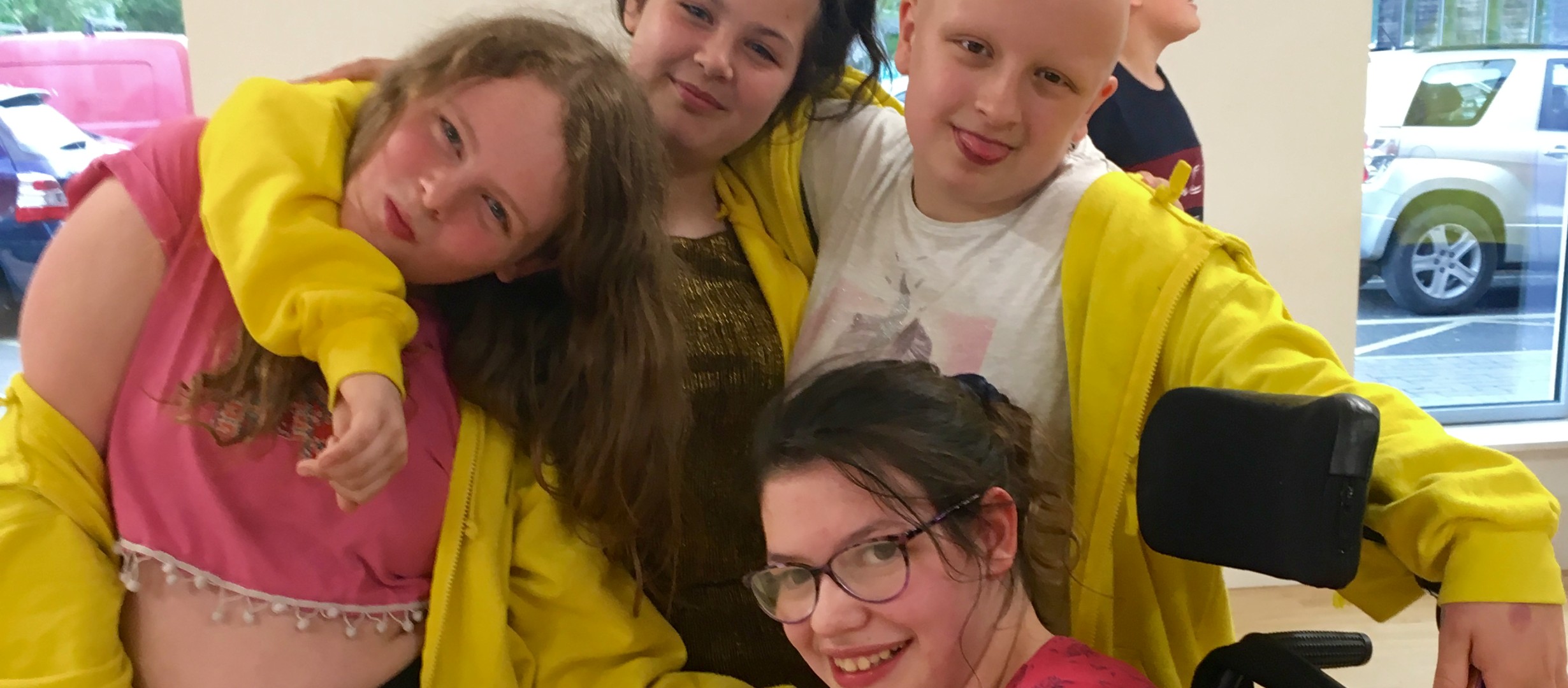 Three young people stood with their arms around each others shoulders. One young person sat in front of the three in a wheelchair. All are smiling at the camera.