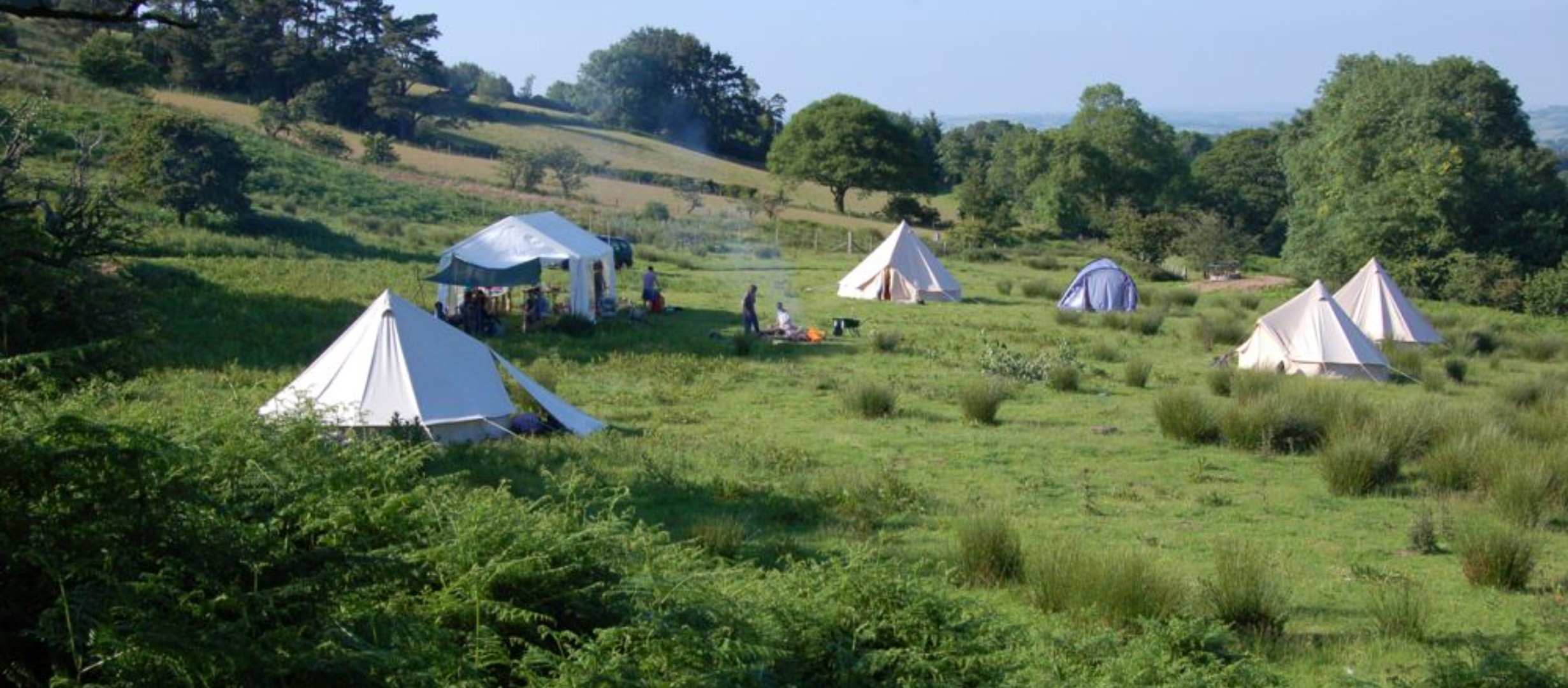 Green field with 6 tents pitched in a circle and a fire in the middle