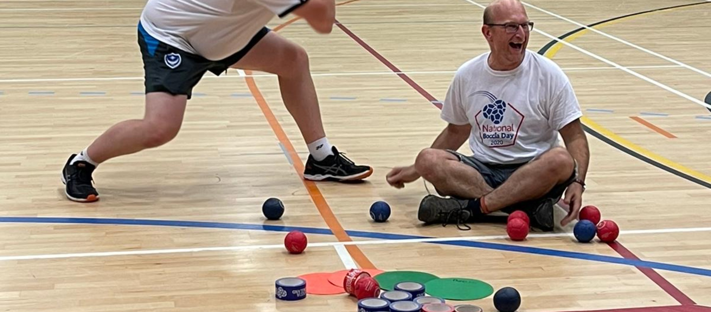 Boccia coach Dave sitting cross legged on the floor surrounded by boccia balls with more balls and equipment in front of him.  The legs of another coach moving toward him are on the left