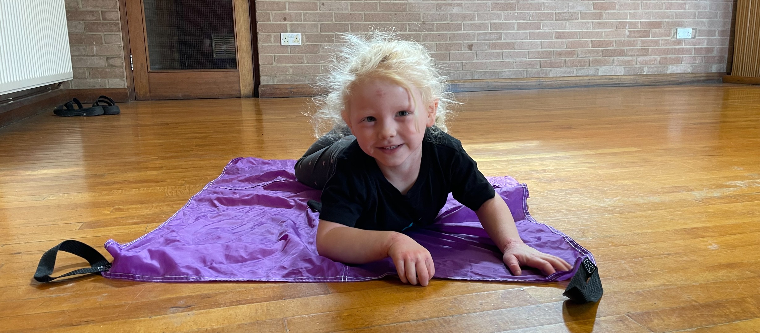 Young child looking at the camera and smiling laying on a purple sheet on a wooden hall. 