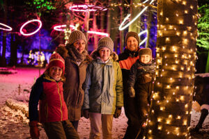 Image shows a family of three children and two adults dressed in warm clothes and standing in woodland surrounded by light installations, including tree trunks wrapped in fairy lights and suspended pink halos