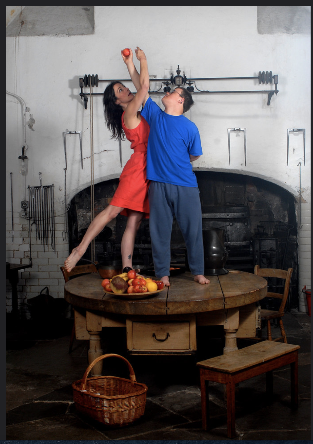 Two dancers stand on a big oak table in a castle kitchen. Their arms are entwined as they both reach up for a red apple. The female dancer has long dark hair. She reaches her right leg away gracefully. She is wearing a short-sleeved to-the-knee bright orange dress. The male dancer has light brown short hair and glasses. He is wearing a royal blue t-shirt and darker blue tracksuit bottoms. They look like a painting from the past.