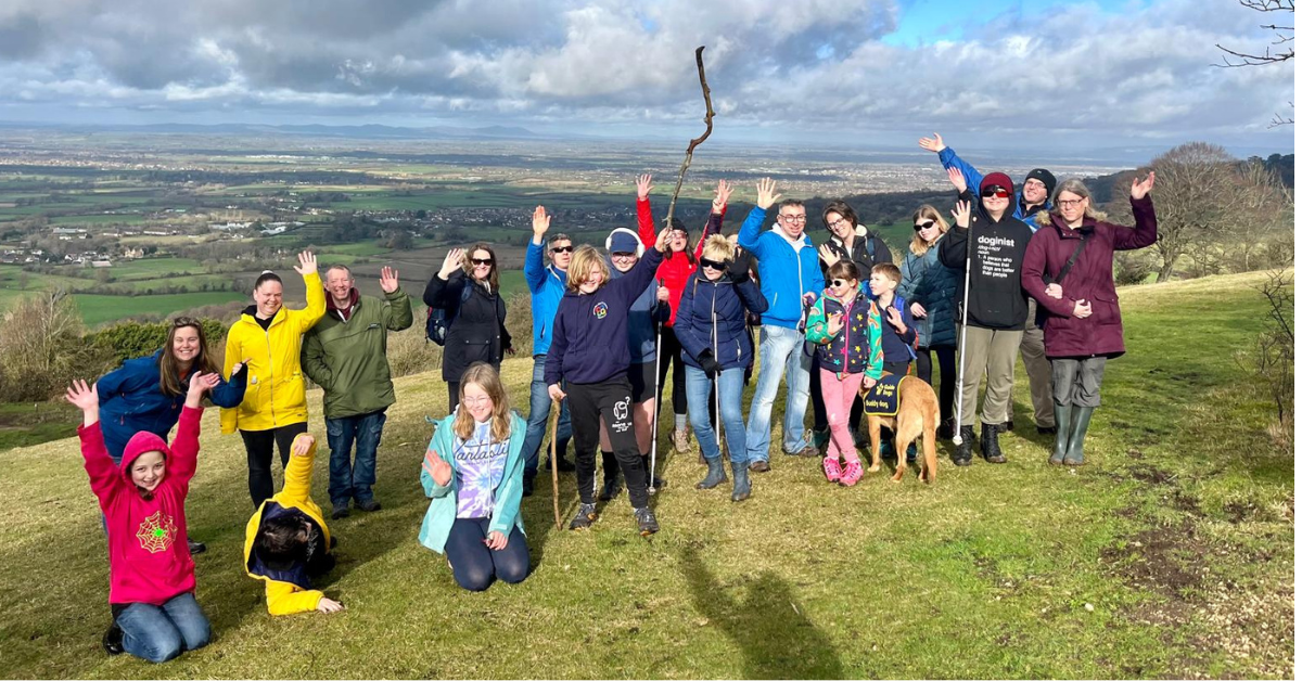 Colour photo of a group of families smiling and waving from the top of Crickley Hill in Gloucester. There is a view of Gloucestershire behind them, with blue sky and clouds.