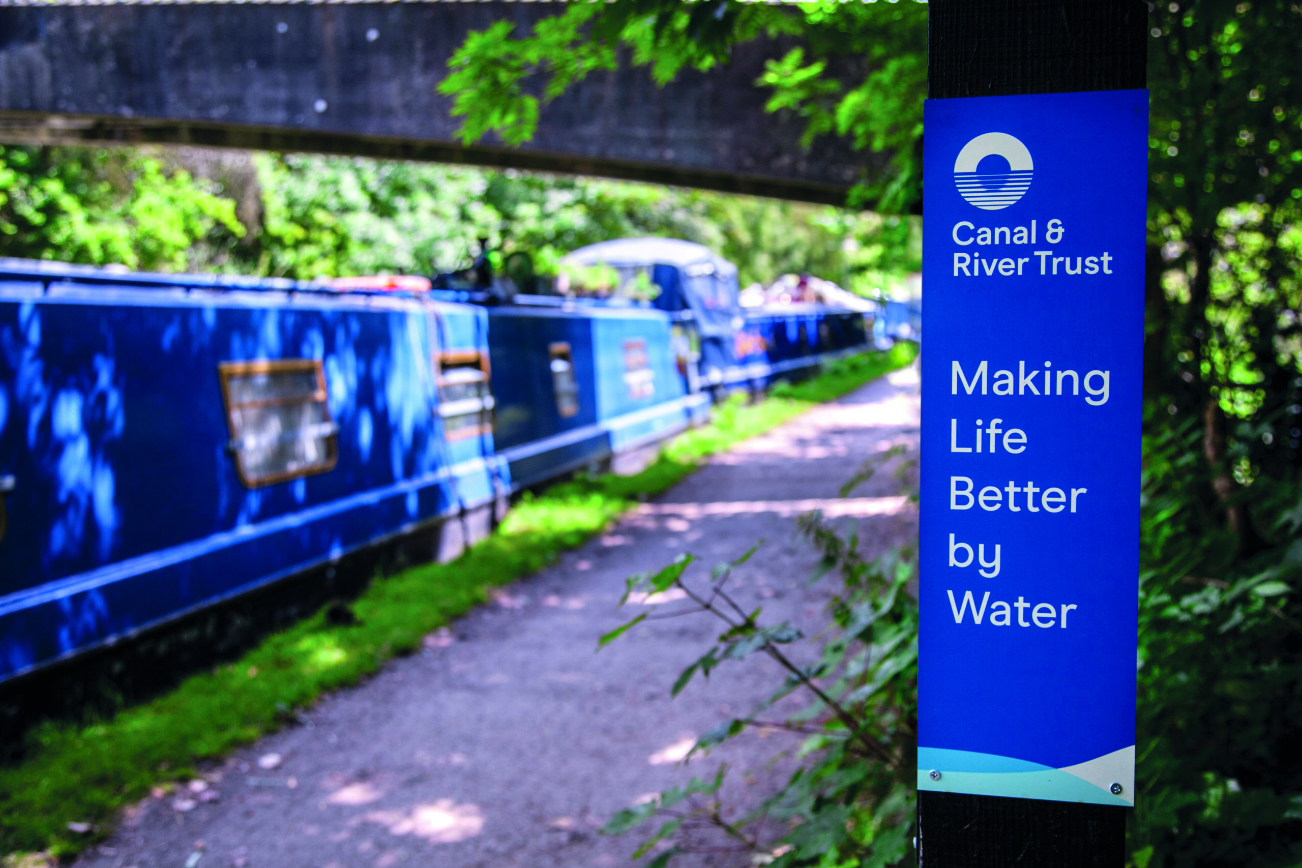 A blue canal boat moored alongside a paved towpath. A sign says 