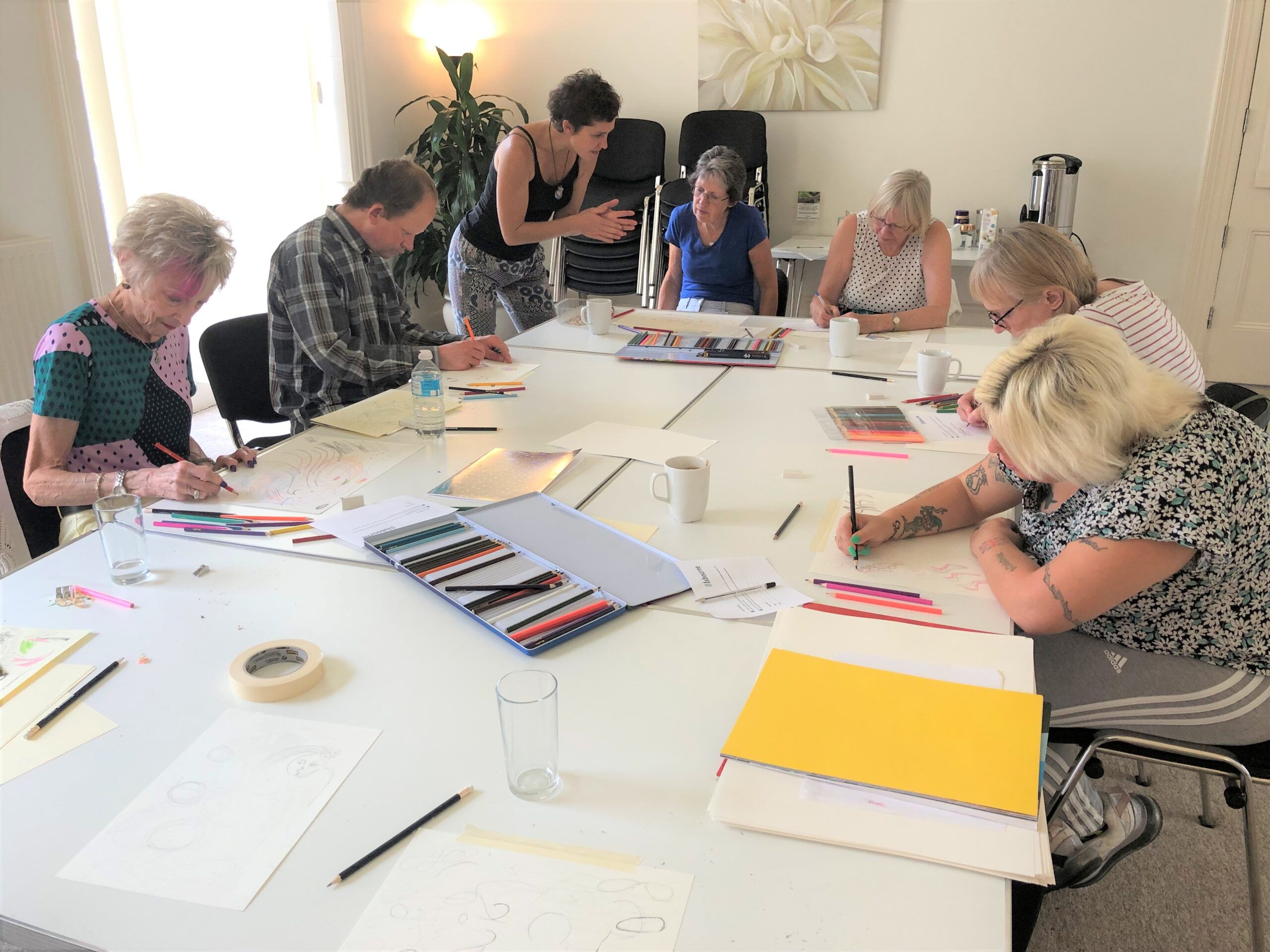 Art for Wellbeing group, with Leah Marshall an affiliated tutor with The Isbourne. A group of community wellbeing attendees are sat round a large table, with art materials, including paper and coloring pencils.
