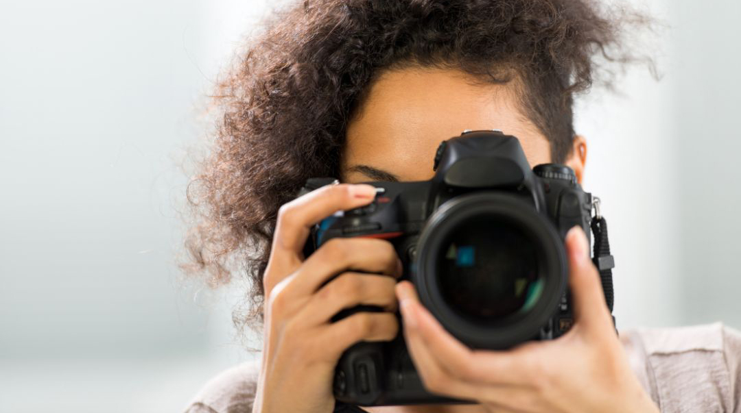 This is a photograph of a lady holding a DSLR camera, her face is hidden by the camera and we see a close up view on the lens