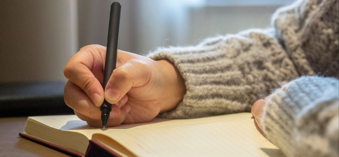 This is a photograph of a person in a cosy grey jumper writing in a journal with a black pen