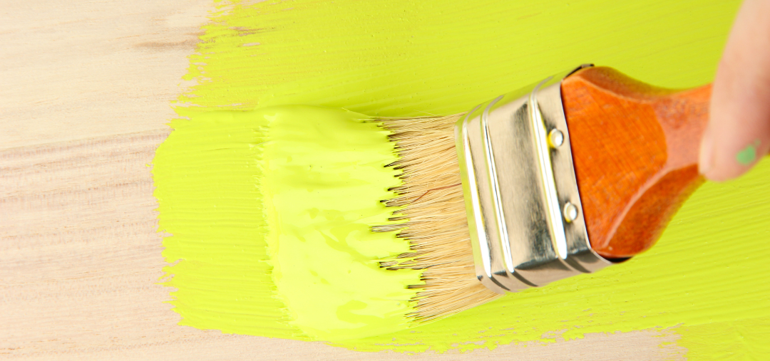 This photograph is of a hand holding a paintbrush with an orange handle. They are using the paintbrush to apply lime green paint onto a wooden surface