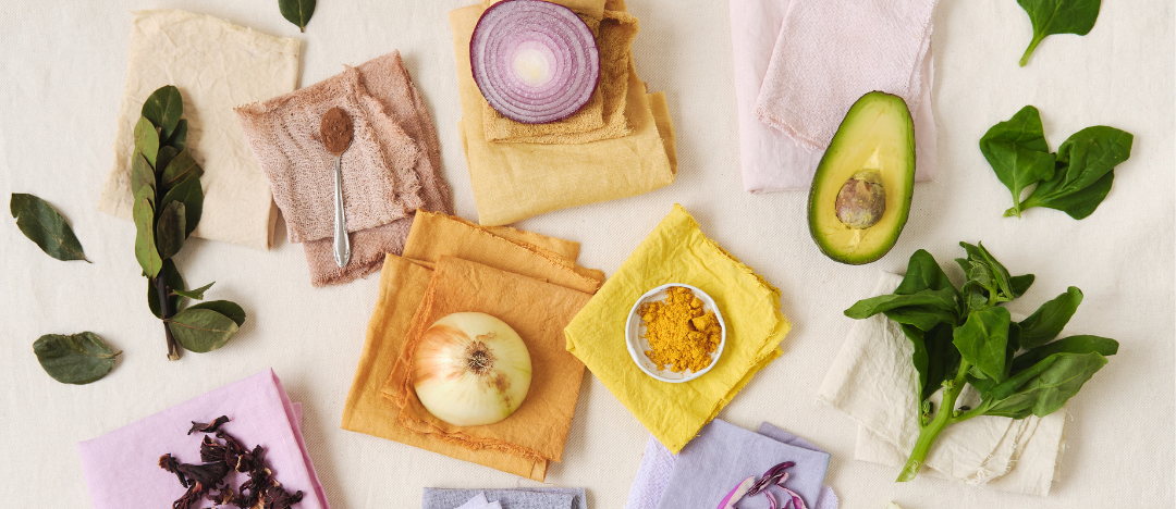 This colourful photograph has various vegetables which can be used to dye fabric. There are onions, turmeric, avocado and bay leaves arranged on squares of cloth in yellow and purple hues. Everything is beautifully displayed on a white table cloth