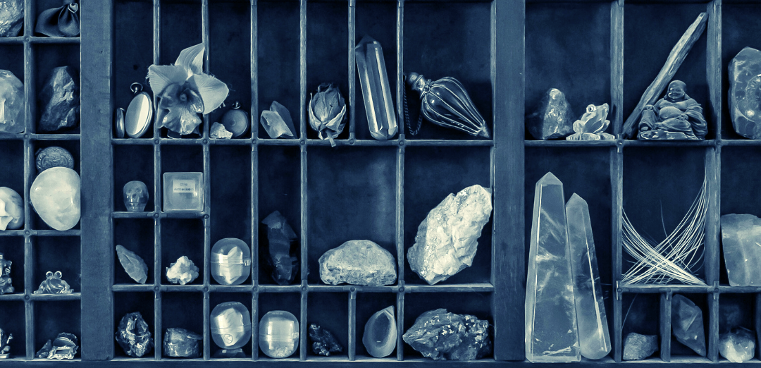 This is a photograph of a cabinet of curiosities. It is made of a wooden case with little compartments full of tiny and interesting objects. You can see large crystals to the right, and a little figure of Budda, and some organic material including small stones. To the left are more stones, dried flowers, a watch, and other mysterious items in little plastic capsuels.