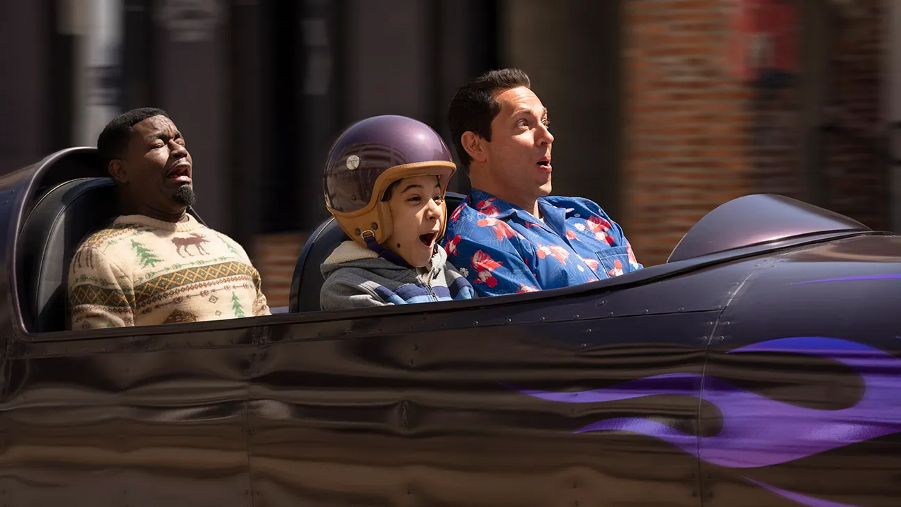 Three people are seated in a black roller coaster car. The person on the left wears a patterned sweater, the middle person wears a helmet, and the person on the right wears a floral shirt. All appear surprised.