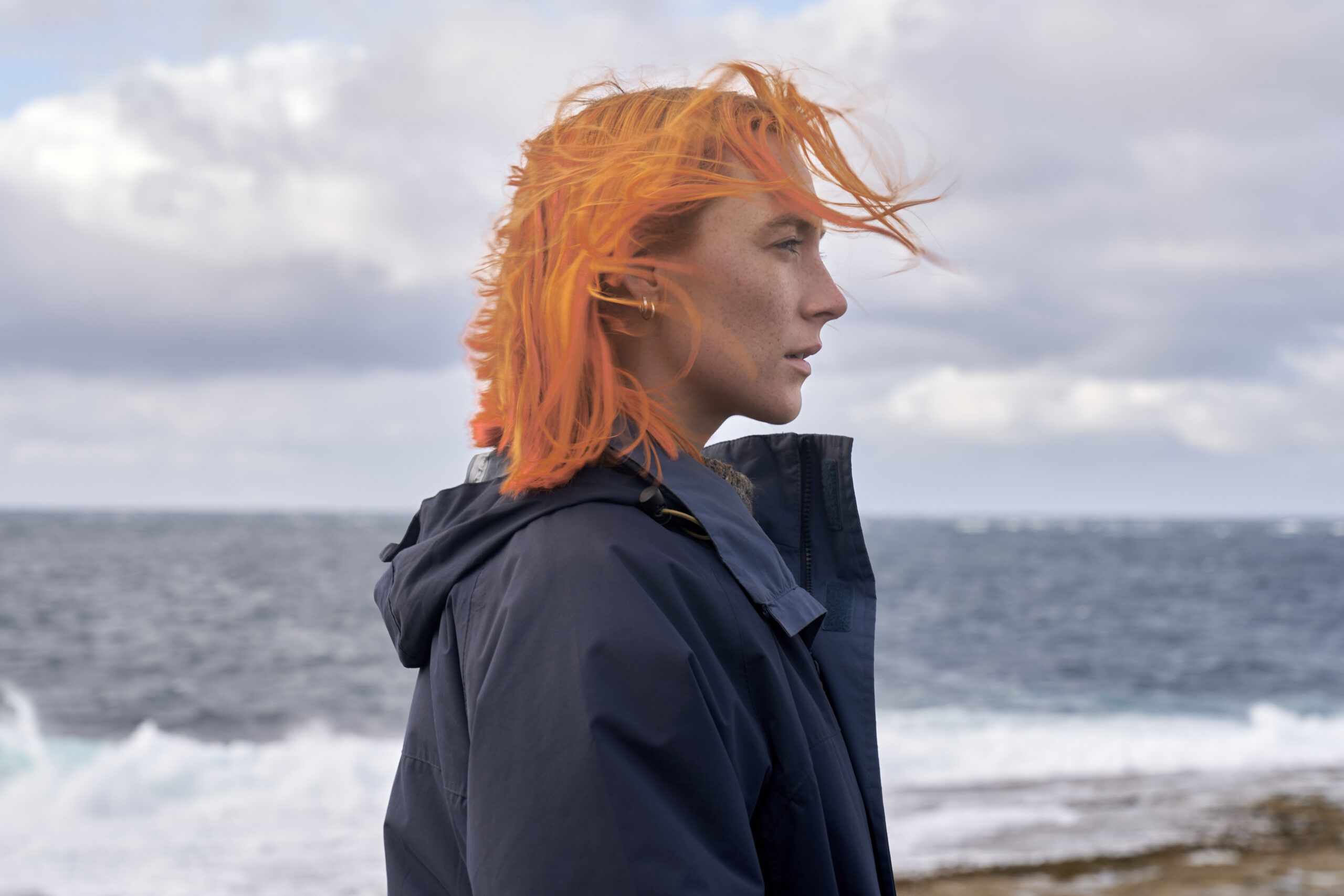 Saoirse Ronan with bright orange hair stood by the sea in a windbreaker