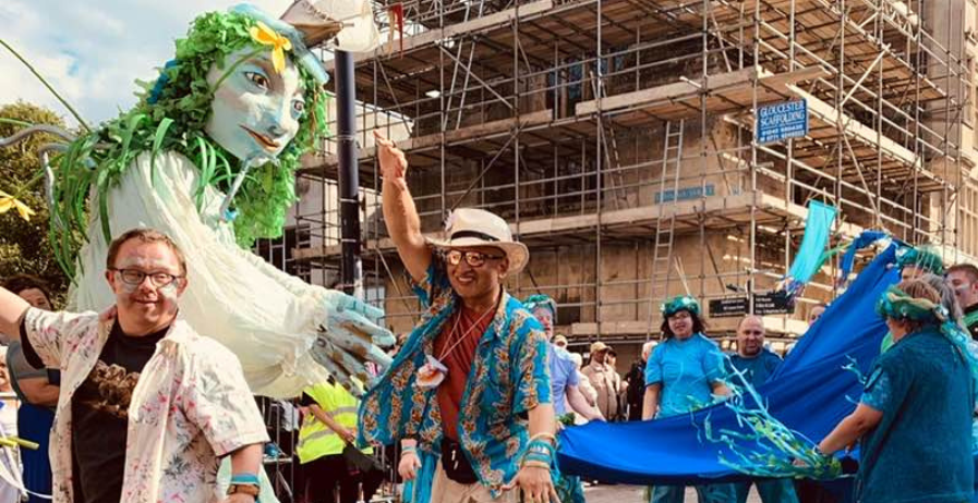 Photograph from Hi Street festival. There are festival performers in the front and a giant puppet with green skin and hair. She is a human depiction of the river Wye.