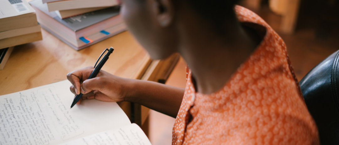 This is a photograph of a woman writing in a journal. She is wearing an orange top