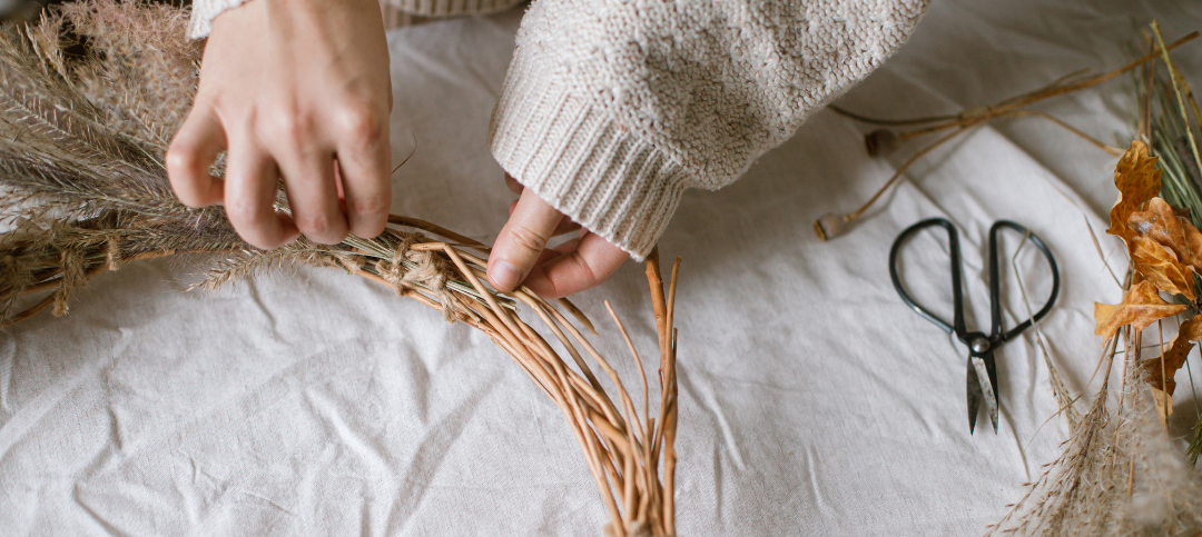 This is a photograph of a woman preparing a beautiful autumn wreath. She is using pampas grass and scissors are on the table. She is working on a white cloth