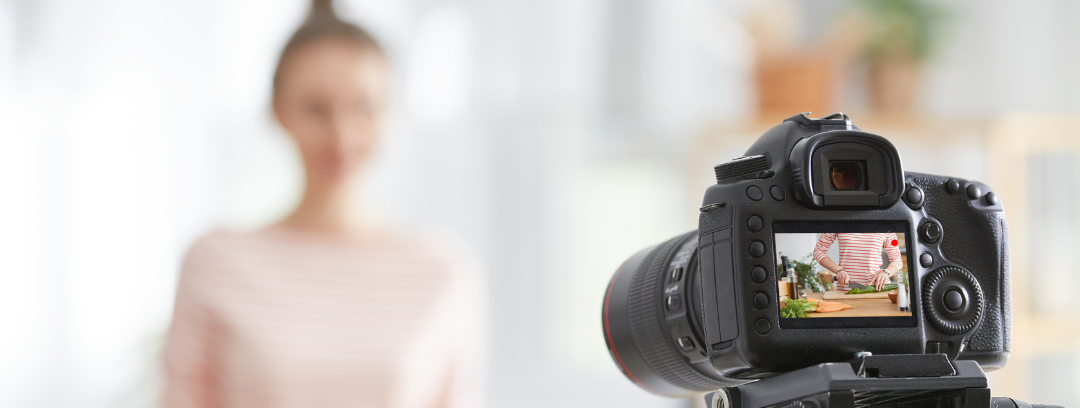 This is a photograph of a woman having her portrait taken. The woman is blurred out but visible in the view finder of the camera. She is preparing food.