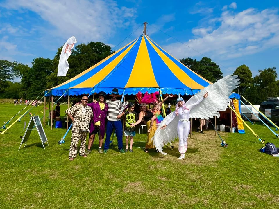 photo of a bright striped big top tent with a lady with a female dressed in a white catsuit with white wings outstretched. Three people are in the foreground, two with bowler style hats. one with juggling clubs and one with a paterned suit. There's a female with a bright pink feathered headress and a child with a smiley face t shirt just behind them. The sky is blue and it looks like a festival