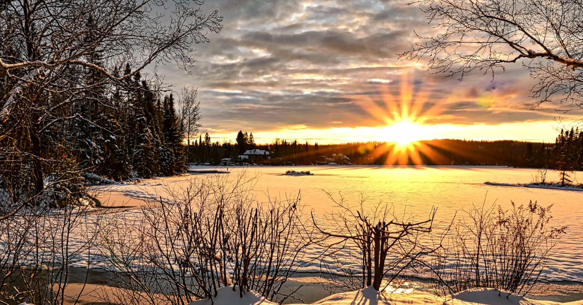 A large field covered in snow. A sunrise in the background and bare tree around the edges.