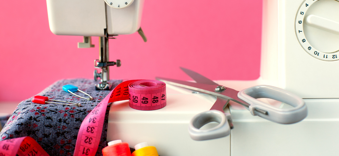 This is a photograph of a sewing machine in front of a bright pink background. There is a pink measuring tape, scissors with a white handle and some different coloured thread.