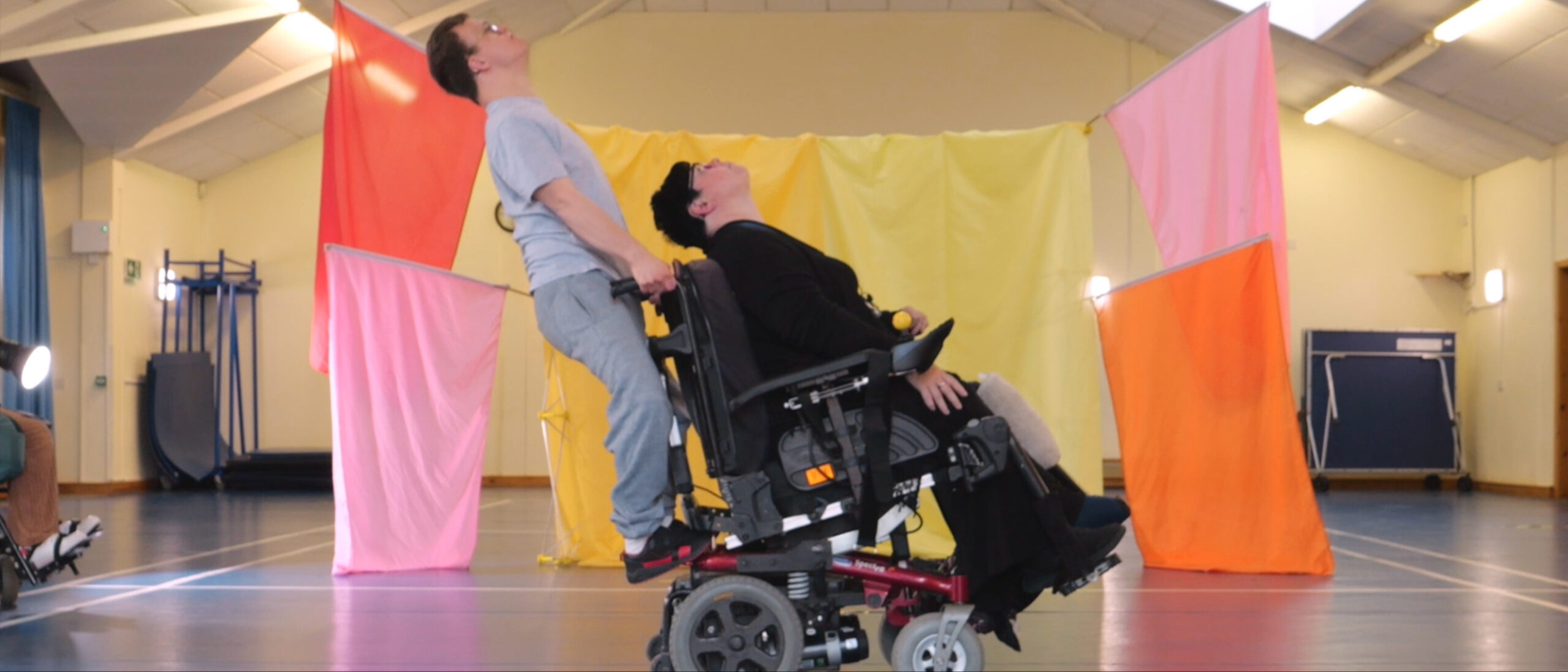 Photograph of two VIA performers . There is a wheelchair user dressed in dark clothes, and a non-wheelchair user in grey. The background is arranged with drapes of fabric in pink, orange and yellow which look like flags