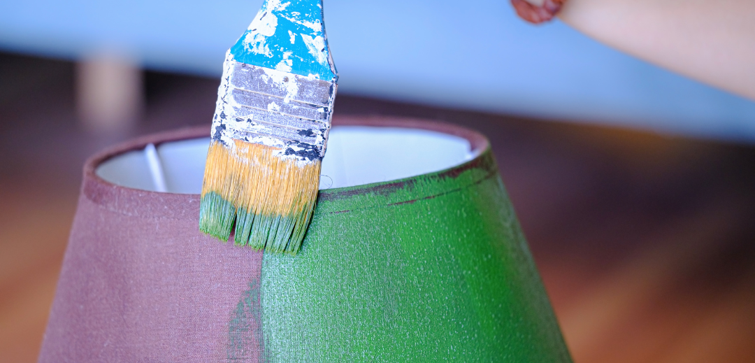 This is a photograph of a woman repainting a lampshade. She is painting over the old purple colour with a new vibrant green