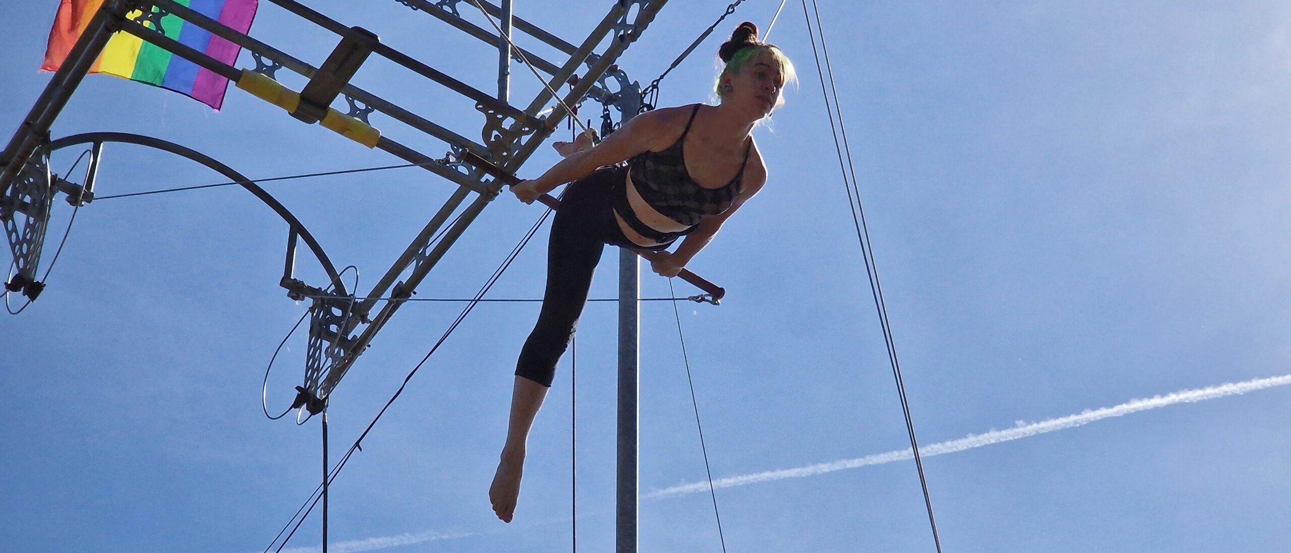 Picture of a flying trapeze artist swinging high in the sky against a blue sky, with a pride flag