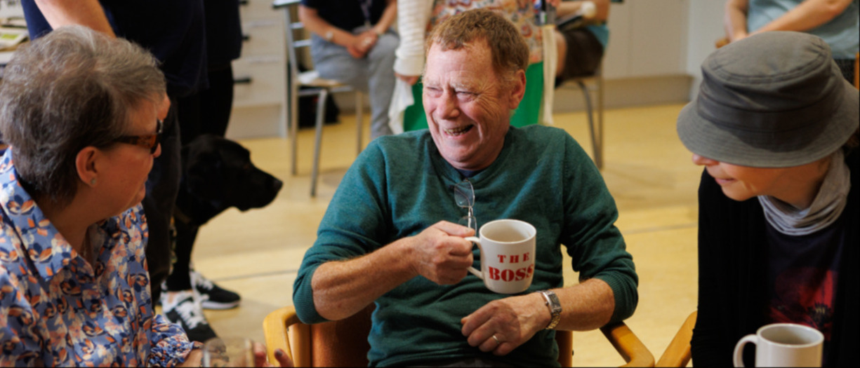 Three people sit at a round table, chatting and laughing whilst drinking tea.