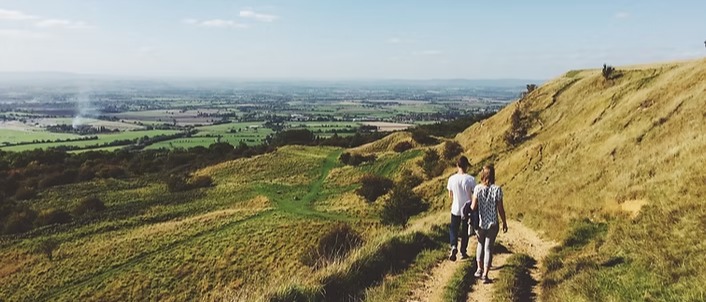 A photo of two people walking in a green landscape on the side of a hill. Their backs are to the camera and we can't see their faces
