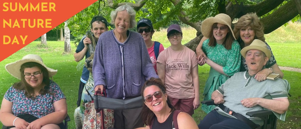 A colour photo of a group of adults and facilitator Louise gathered in a park. Everyone is smiling.