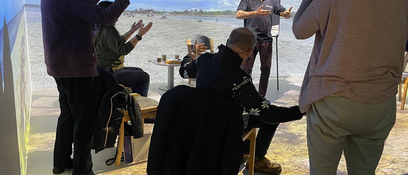 The image is of a group of people in a circle in the Oakley interactive space. On the walls of the room is a digital image of a beach, with blue skies.