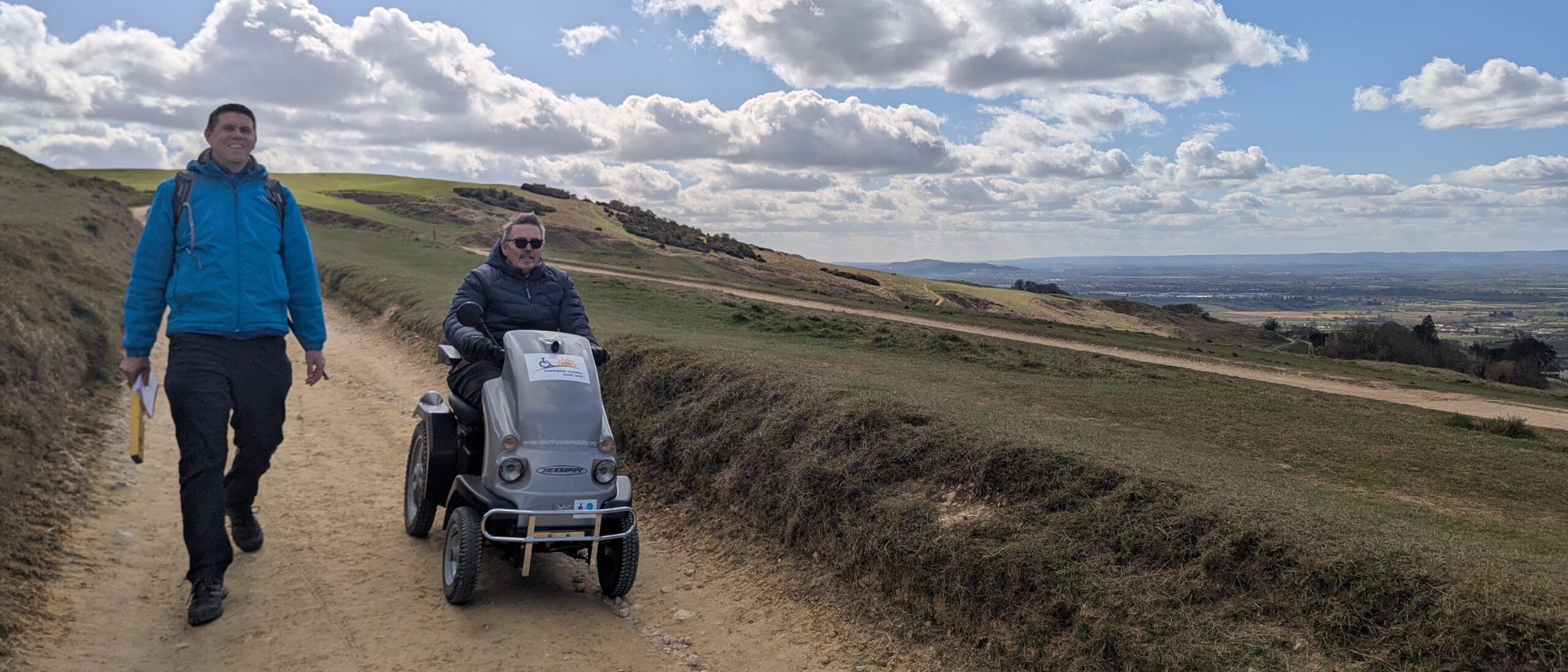 Two men on a path on Cleeve Common. One is walking and wearing a blue coat. the other is using an off-road mobility scooter.